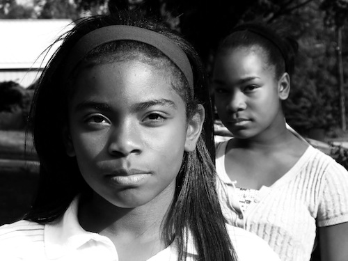 Fotografia de duas meninas negras, à frente uma menina de cabelo liso com faixa no cabelo e blusa branca, mais atrás uma menina de blusa branca e cabelo preso
